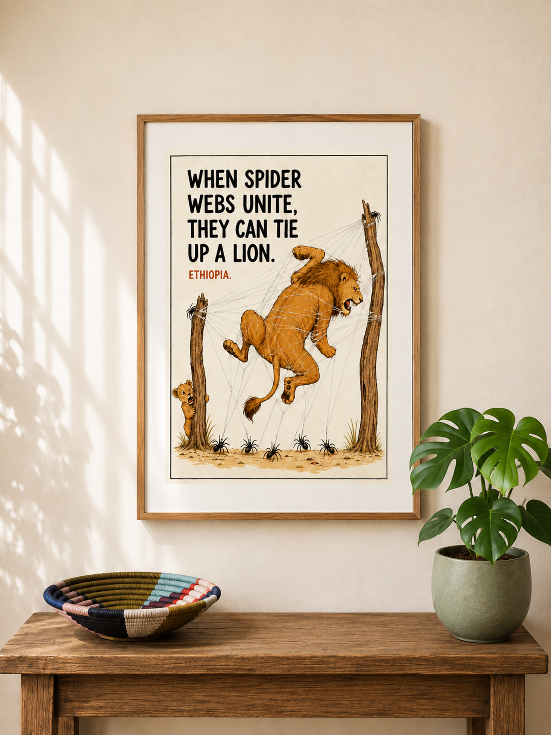 Framed illustrated art print from Ethiopia — a roaring lion tangled and suspended in giant spider webs stretched between trees, with spiders watching from below. Proverb: When spider webs unite, they can tie up a lion. From Mopti's African Proverbs Series.