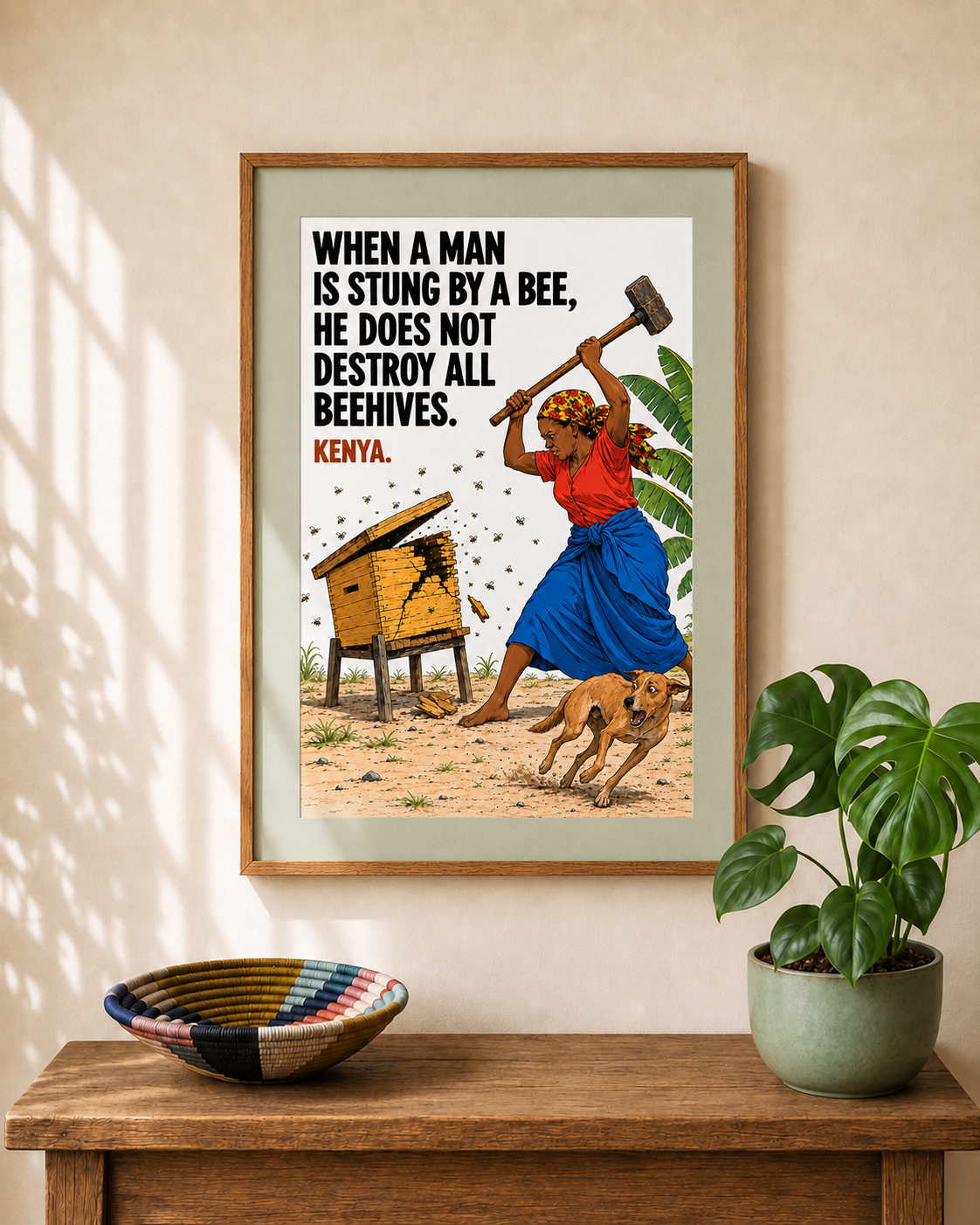 Framed wall art hanging on a beige wall above a wooden console table, featuring a Kenyan quote about a bee sting and an illustrated scene of a woman with an axe and a beekeeper's hatbox. A potted plant and a patterned bowl sit on the table.