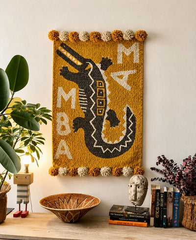 Handmade wool wall hanging featuring a bold crocodile and MAMBA lettering on a mustard ground, with pompom edging – Mopti Living