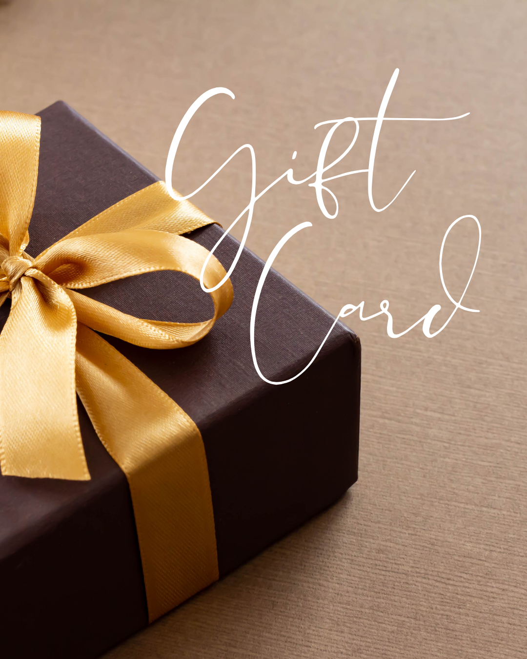 Dark gift box with gold satin ribbon and bow on a warm sandy surface — Mopti Living Gift Card