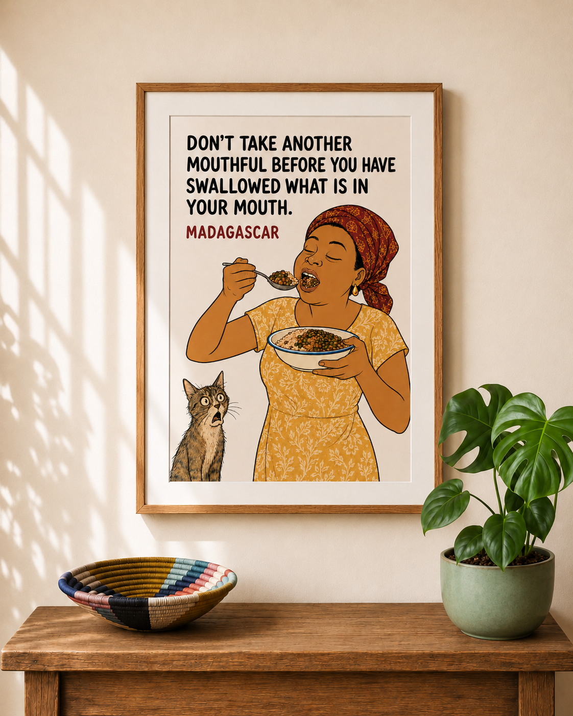 Framed illustrated art print from Madagascar — a woman in a yellow dress eats enthusiastically from a bowl while a wide-eyed cat watches from below. Proverb: Don't take another mouthful before you have swallowed what is in your mouth. From Mopti's African Proverbs Series.